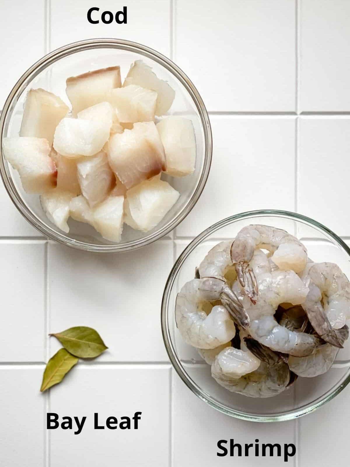 Recipe ingredients in bowls on white board including cod fish shrimp and bay leaf.