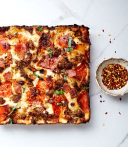 Cooked pizza with bowl of red crushed pepper on a white marble top.