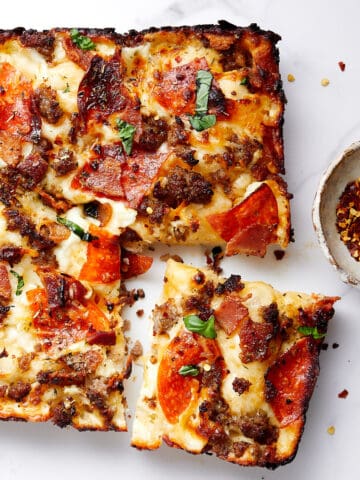 Cooked pizza with cut slice with bowl of red crushed pepper on a white marble top.