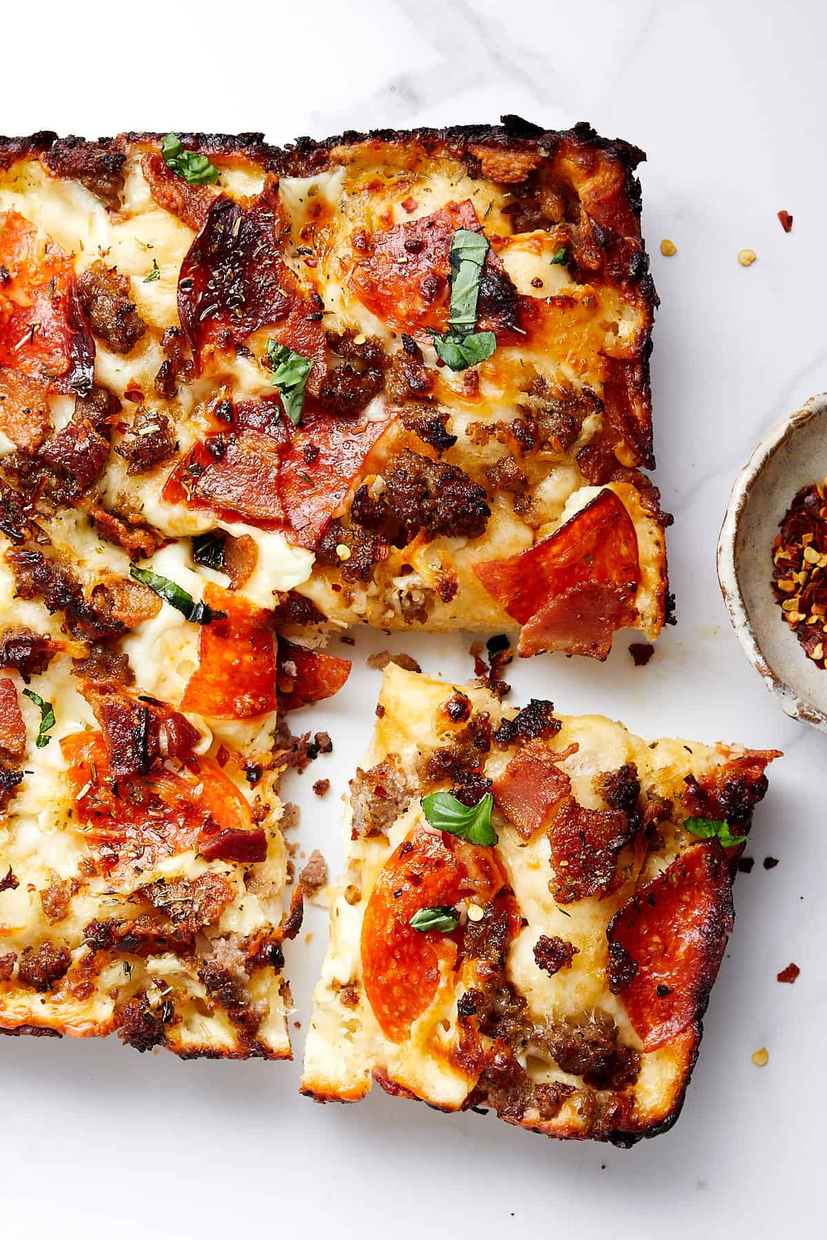 Cooked pizza with cut slice with bowl of red crushed pepper on a white marble top.