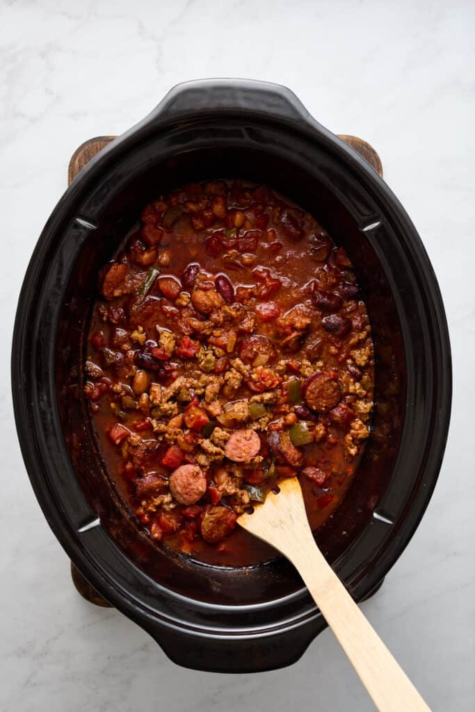 Cooked chili in a black slow cooker vessel with wooden spoon.