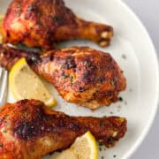 Cooked tandoori chicken leges on white plate with lemon slices and fork.