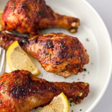 Cooked tandoori chicken leges on white plate with lemon slices and fork.