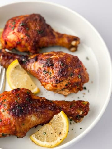 Cooked tandoori chicken leges on white plate with lemon slices and fork.
