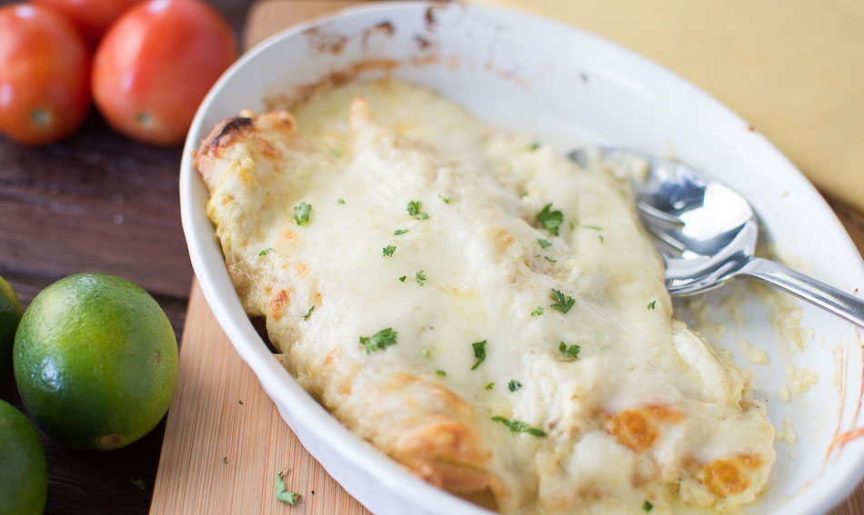 Side view of the White Sauce Enchiladas with Salsa Verde in a white tray with limes and tomatoes on the side